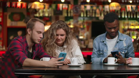 Eine-Gruppe-Von-Freunden-Sitzt-In-Einer-Bar,-Trinkt-Kaffee-Und-Unterhält-Sich,-Während-Sie-Auf-Den-Bildschirm-Eines-Smartphones-Schauen.